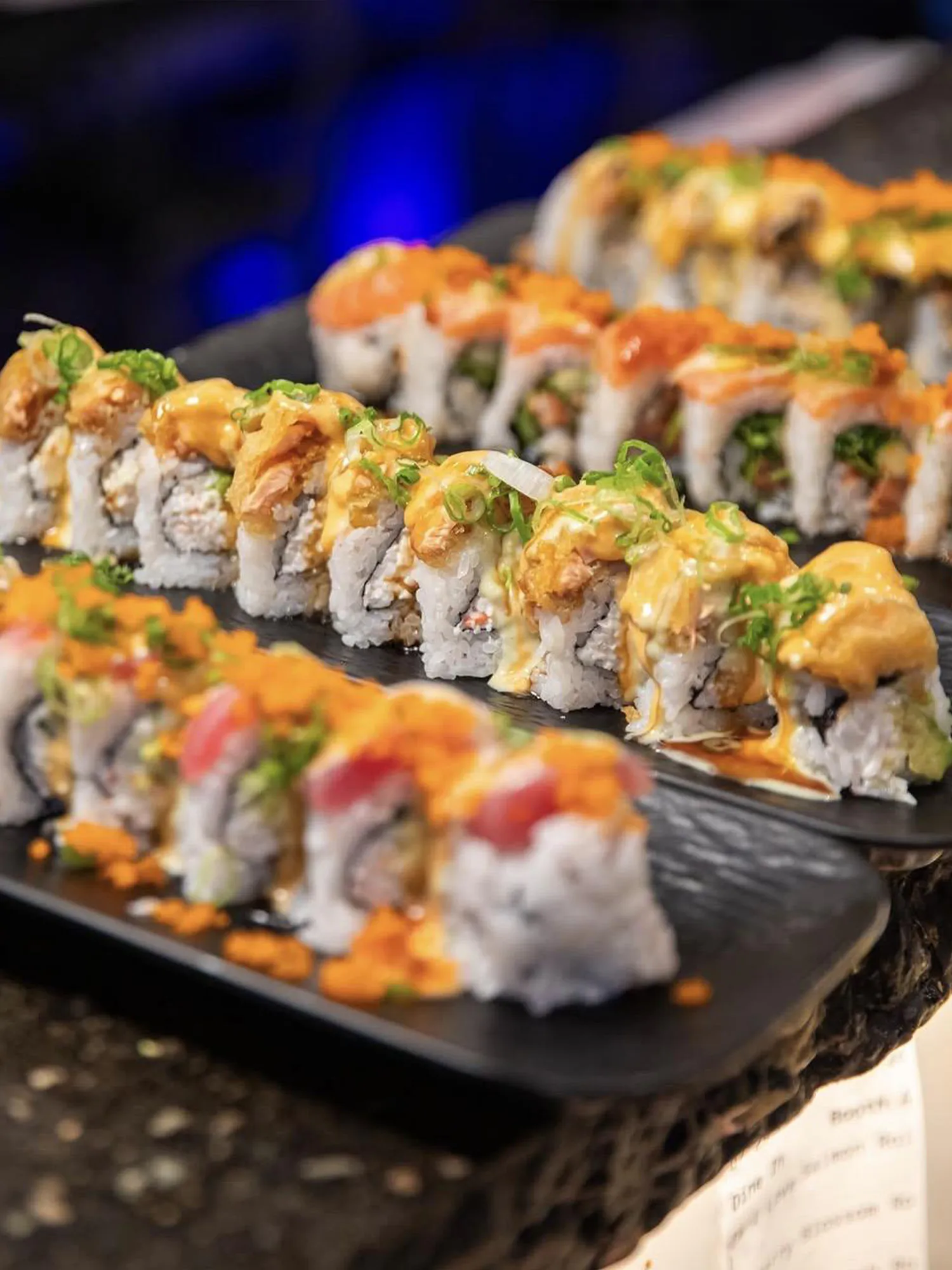 Assorted sushi rolls topped with spicy mayo, green onions, and roe on black plates.