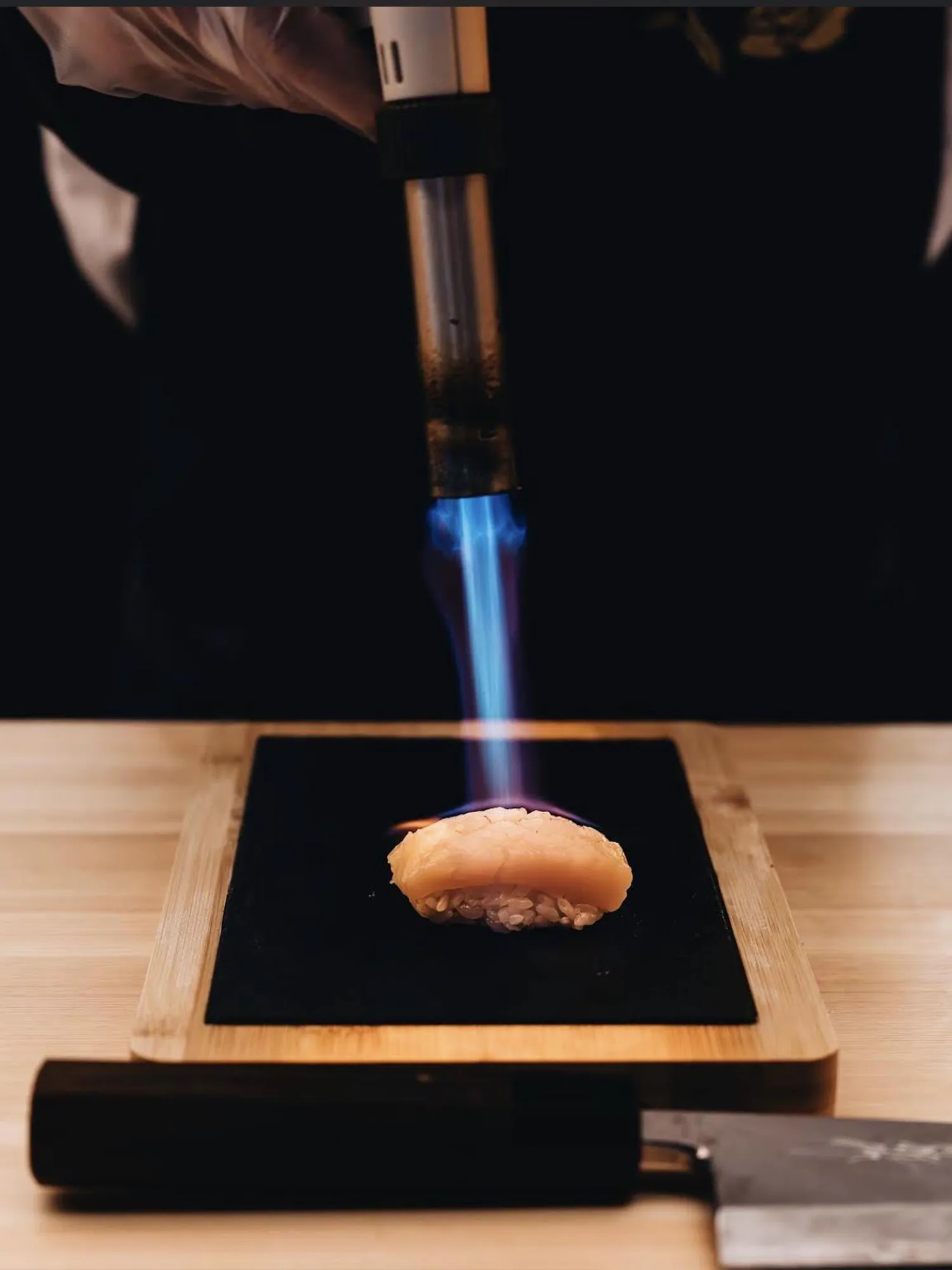 Chef using a blowtorch to sear a piece of nigiri sushi on a wooden board.